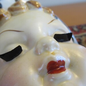 Puede incluir: Primer plano de una cabeza de figura de cer&aacute;mica vintage. La cara es de color crema p&aacute;lido con p&aacute;rpados negros, cejas marrones y labios rojos. El cabello est&aacute; peinado en peque&ntilde;os rizos redondeados con detalles marrones.