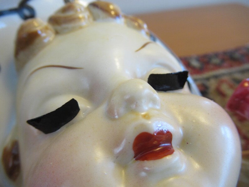 Puede incluir: Primer plano de una cabeza de figura de cer&aacute;mica vintage. La cara es de color crema p&aacute;lido con p&aacute;rpados negros, cejas marrones y labios rojos. El cabello est&aacute; peinado en peque&ntilde;os rizos redondeados con detalles marrones.