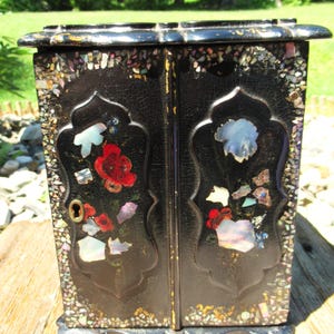 May include: A black jewelry box with two doors featuring inlaid mother-of-pearl floral designs and a mosaic border. The surface has a crackled finish, and the left door includes a brass keyhole escutcheon. The box has a scalloped base and top.