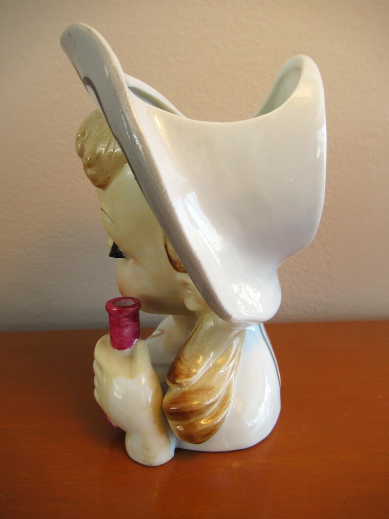 Puede incluir: Jarra de cer&aacute;mica vintage con forma de mujer sosteniendo una botella roja. La jarra es principalmente blanca con detalles en beige, marr&oacute;n y negro. La mujer lleva un sombrero blanco grande. Probablemente un art&iacute;culo decorativo.