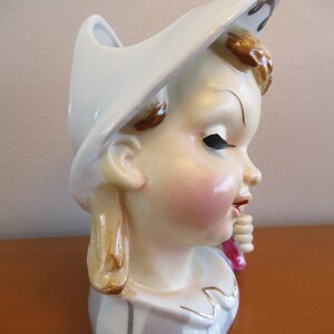 Puede incluir: Jarr&oacute;n de cer&aacute;mica vintage con forma de cabeza de ni&ntilde;a, con un sombrero blanco. El jarr&oacute;n tiene una cara pintada con mejillas rosadas, cabello casta&ntilde;o y una camisa a rayas. La ni&ntilde;a sostiene un objeto rosa.