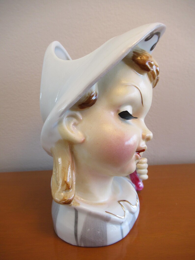 Puede incluir: Jarr&oacute;n de cer&aacute;mica vintage con forma de cabeza de ni&ntilde;a, con un sombrero blanco. El jarr&oacute;n tiene una cara pintada con mejillas rosadas, cabello casta&ntilde;o y una camisa a rayas. La ni&ntilde;a sostiene un objeto rosa.