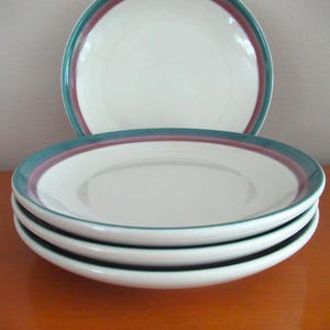 May include: A stack of four white ceramic plates with a teal and burgundy border. One additional plate is visible in the background. The plates are stacked on top of each other, with the top plate slightly angled. The plates are on a wooden surface.
