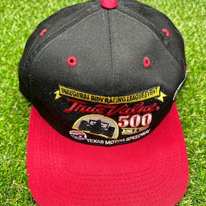 Puede incluir: Gorra de béisbol negra y roja con un parche bordado amarillo y rojo que dice "Inaugural Indy Racing League Event True Value 500 June 2, 1997 Texas Motor Speedway".