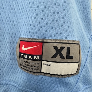 K&ouml;nnte beinhalten: Ein hellblaues Sporttrikot mit einem Nike-Logo und dem Text "TEAM" und "ENGINEERED TO THE EXACT SPECIFICATIONS OF CHAMPIONSHIP ATHLETES". Das Trikot hat ein Gr&ouml;&szlig;enetikett mit der Aufschrift "XL" und "NAME/#".