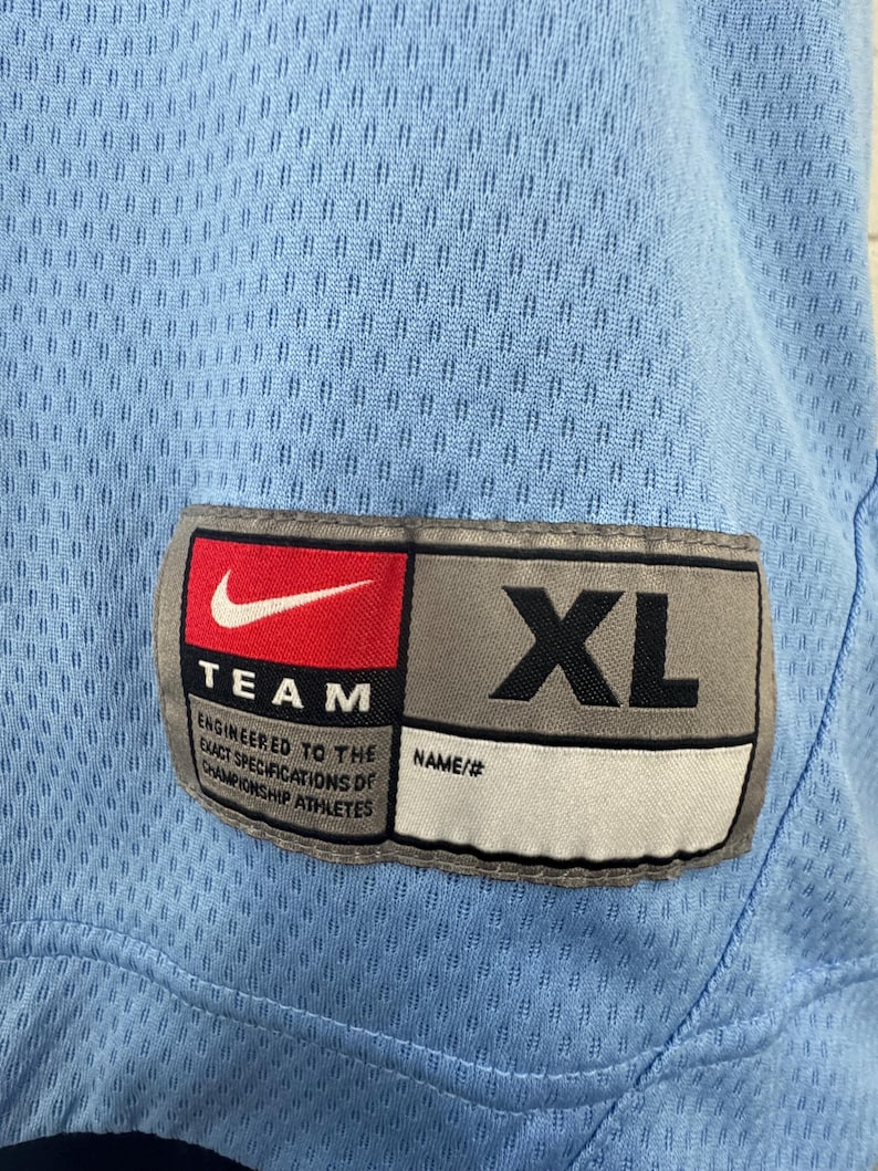 K&ouml;nnte beinhalten: Ein hellblaues Sporttrikot mit einem Nike-Logo und dem Text "TEAM" und "ENGINEERED TO THE EXACT SPECIFICATIONS OF CHAMPIONSHIP ATHLETES". Das Trikot hat ein Gr&ouml;&szlig;enetikett mit der Aufschrift "XL" und "NAME/#".
