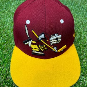 Puede incluir: Una gorra de béisbol de color granate y dorado con una mascota de dibujos animados que lleva un uniforme de hockey. La mascota sostiene un palo de hockey y un disco. La gorra tiene una visera plana.
