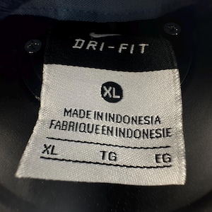 K&ouml;nnte beinhalten: Ein wei&szlig;es Etikett mit schwarzem Text, auf dem "Dri-Fit" und "Made in Indonesia Fabrique en Indonesie" steht. Das Etikett hat auch ein Gr&ouml;&szlig;enetikett mit der Aufschrift "XL" und drei leere Felder mit den Bezeichnungen "XL", "TG" und "EG".