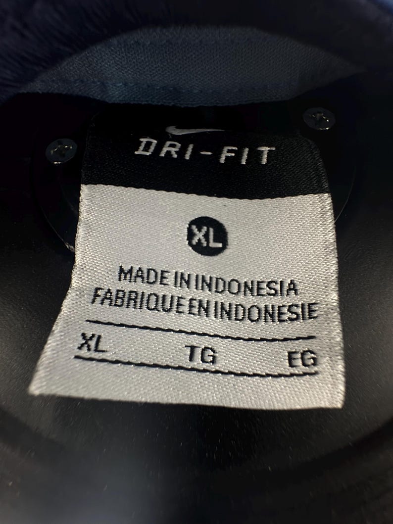 K&ouml;nnte beinhalten: Ein wei&szlig;es Etikett mit schwarzem Text, auf dem "Dri-Fit" und "Made in Indonesia Fabrique en Indonesie" steht. Das Etikett hat auch ein Gr&ouml;&szlig;enetikett mit der Aufschrift "XL" und drei leere Felder mit den Bezeichnungen "XL", "TG" und "EG".