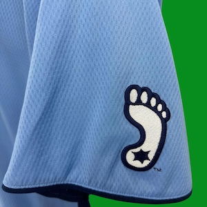 K&ouml;nnte beinhalten: Hellblaues Trikot mit einem wei&szlig;en und marineblauen Fu&szlig;abdruck-Logo auf dem &Auml;rmel. Das Logo hat einen Stern im Fu&szlig;abdruck. Der Text "TM" befindet sich unter dem Logo.