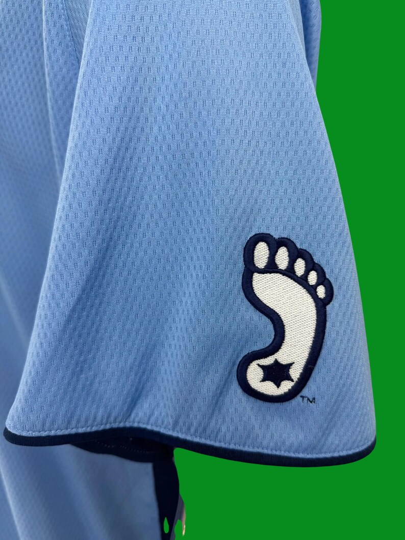 K&ouml;nnte beinhalten: Hellblaues Trikot mit einem wei&szlig;en und marineblauen Fu&szlig;abdruck-Logo auf dem &Auml;rmel. Das Logo hat einen Stern im Fu&szlig;abdruck. Der Text "TM" befindet sich unter dem Logo.