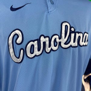 K&ouml;nnte beinhalten: Ein hellblaues Baseballtrikot mit einem wei&szlig;en Nike-Swoosh-Logo auf der linken Brust und dem Wort "Carolina" in wei&szlig;en und dunkelblauen Buchstaben auf der Vorderseite.