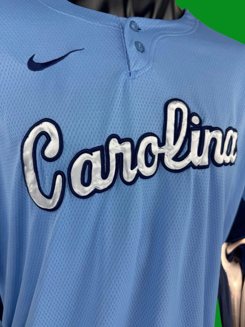 K&ouml;nnte beinhalten: Ein hellblaues Baseballtrikot mit einem wei&szlig;en Nike-Swoosh-Logo auf der linken Brust und dem Wort "Carolina" in wei&szlig;en und dunkelblauen Buchstaben auf der Vorderseite.
