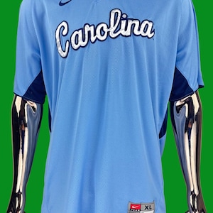 K&ouml;nnte beinhalten: Ein hellblaues Baseballtrikot mit dem Wort "Carolina" in wei&szlig;er und dunkelblauer Schrift auf der Vorderseite. Das Trikot hat ein dunkelblaues Nike-Swoosh-Logo auf der linken Brust und ein dunkelblaues und wei&szlig;es Nike-Teamlogo mit der Gr&ouml;&szlig;e "XL" unten rechts.