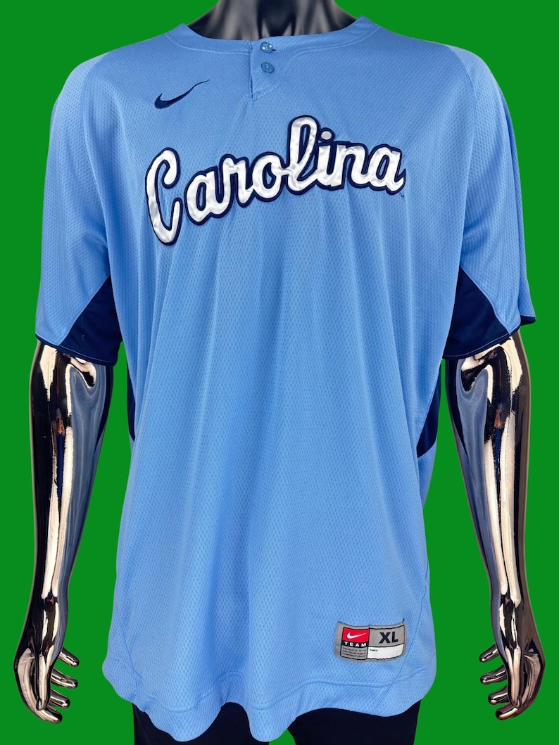 K&ouml;nnte beinhalten: Ein hellblaues Baseballtrikot mit dem Wort "Carolina" in wei&szlig;er und dunkelblauer Schrift auf der Vorderseite. Das Trikot hat ein dunkelblaues Nike-Swoosh-Logo auf der linken Brust und ein dunkelblaues und wei&szlig;es Nike-Teamlogo mit der Gr&ouml;&szlig;e "XL" unten rechts.