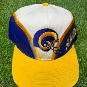 May include: A white, blue and yellow baseball cap with a Rams logo embroidered on the front. The logo is a blue and yellow ram's head wearing a helmet. The cap has a yellow brim.