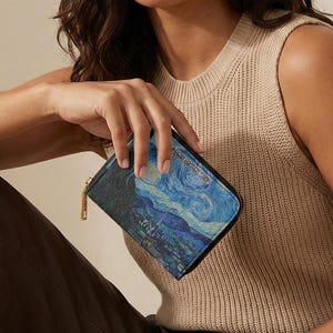 May include: A rectangular zip-around wallet featuring a printed reproduction of Vincent van Gogh's The Starry Night painting on the front panel. The wallet has a dark textured leather-like side panel and a gold-toned metal zipper closure.