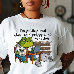 May include: White t-shirt featuring a cartoon frog working on a laptop with the text "I'm getting real close to a grippy sock vacation." The frog is wearing a tie and holding a coffee mug. A cat is lying next to the desk.