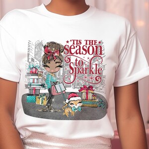 May include: White t-shirt with a festive graphic of a cartoon character, gifts, and a dog in a Santa hat. The text "'Tis the season to Sparkle" is displayed in red and white. The t-shirt is worn by a person with pink hair, perfect for the holiday season.