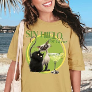 May include: Mustard yellow t-shirt featuring a cartoon illustration of two cats and a margarita. The text "SIN HIELO, por favor" is above the image. The design is inside a green circle.