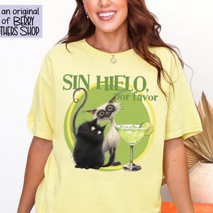 May include: Yellow t-shirt featuring a graphic of two cats and a margarita glass. The text "SIN HIELO, por favor" is above the image. The shirt has a relaxed fit and short sleeves. The design is an original design of BERRY BROTHERS SHOP.