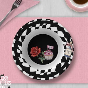 May include: A black and white checkered bowl with a black interior featuring a red rose and cupcake illustration. The bowl is on a pink placemat with a fork and napkin. A teacup and saucer are in the upper right corner. The bowl has the text "Drink Me" and "Eat Me".
