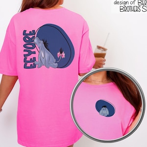 May include: A bright pink t-shirt featuring a cartoon Eeyore design. The design includes a blue oval, the name "Eeyore" in purple, and a small Eeyore illustration with a pink bow. The text "This is an original design of BERRY BROTHERS SHOP" is also visible.