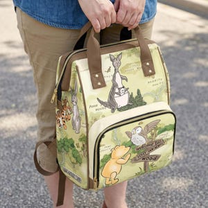 May include: A beige backpack featuring a printed map of the Hundred Acre Wood with illustrations of Winnie the Pooh, Tigger, Rabbit, Kanga, Roo, and Owl. The bag has brown straps, gold-tone zippers, and a sign reading Welcome to 100 Aker Wood.