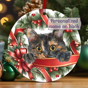 May include: A round ceramic Christmas ornament featuring a watercolor illustration of a tortoiseshell cat peeking over a red ribbon. The cat is surrounded by a festive wreath of green pine needles, holly berries, and red ribbons. Text reads: Personalized name on back.