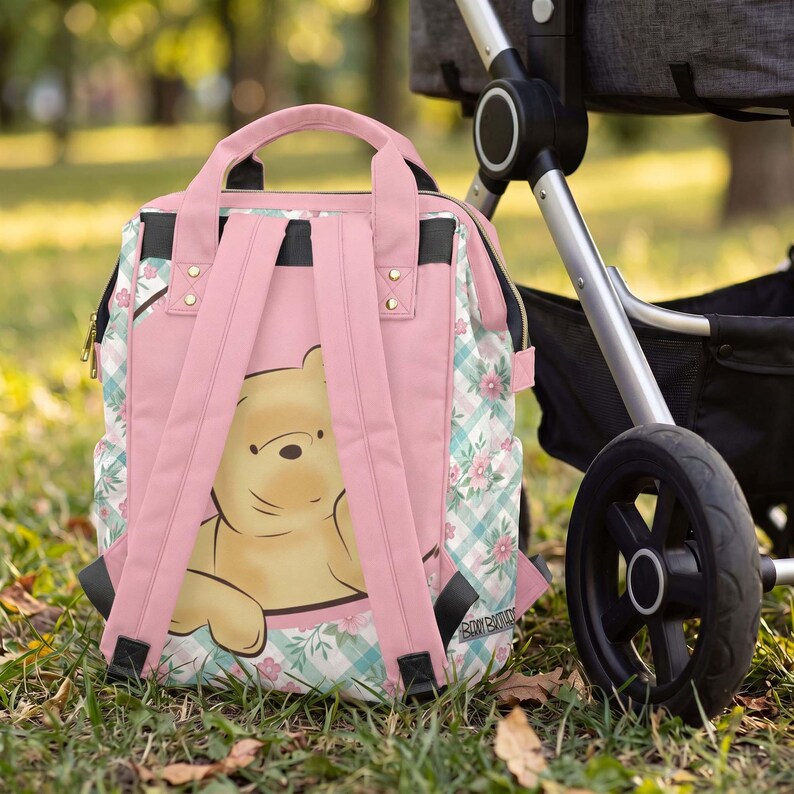 May include: A pink backpack featuring a graphic of Winnie the Pooh on the front panel. The bag has light pink shoulder straps, top handles, and side panels decorated with a light blue and white gingham pattern accented by small pink flowers.