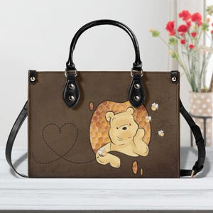 May include: A brown rectangular handbag with black handles and a shoulder strap. The front features a graphic of a light yellow bear character emerging from a honeycomb pattern, surrounded by small flying bees and a dashed heart-shaped flight path.