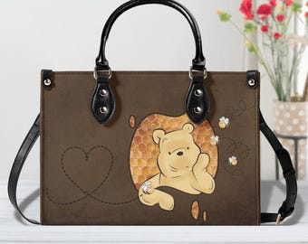 May include: A brown rectangular handbag featuring a graphic of a yellow bear character emerging from a honeycomb pattern. The bag has black handles, black side straps, and small bee illustrations with dashed flight paths on the front.