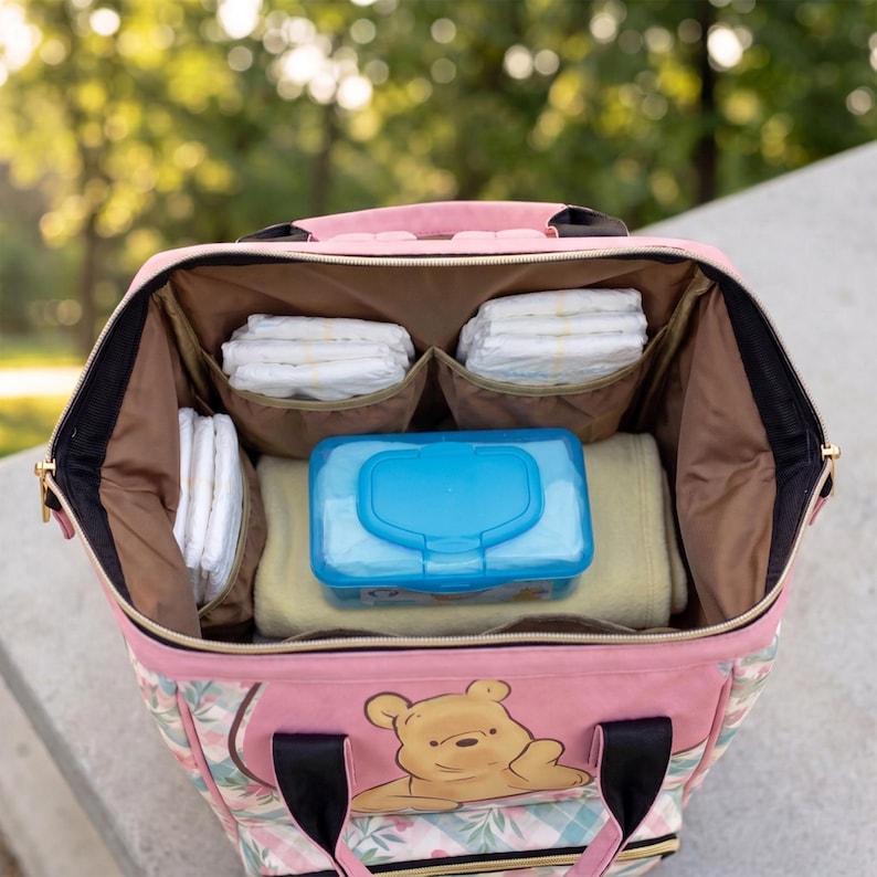 May include: A pink diaper bag with a Winnie the Pooh graphic on the front. The interior is lined with brown fabric and contains stacks of white diapers in side pockets, a light yellow blanket, and a bright blue plastic baby wipe container.