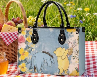 May include: A rectangular handbag featuring a light blue background with a printed illustration of Winnie the Pooh, Piglet, and Eeyore surrounded by yellow and pink flowers. The bag has two black handles, black trim, and gold-tone metal hardware.
