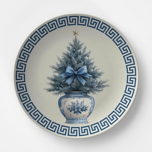 Puede incluir: Un plato redondo de cer&aacute;mica con fondo color crema y un borde azul con dise&ntilde;o de greca griega. En el centro, un &aacute;rbol de Navidad azul dentro de un jarr&oacute;n con motivos azules y blancos, decorado con un lazo azul claro y una estrella dorada.