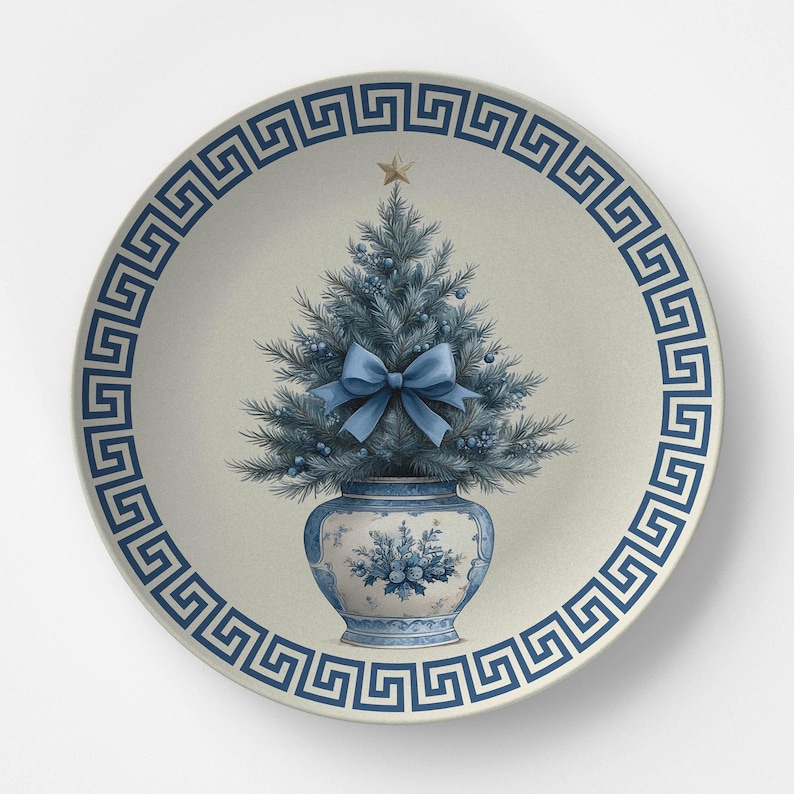 Puede incluir: Un plato redondo de cer&aacute;mica con fondo color crema y un borde azul con dise&ntilde;o de greca griega. En el centro, un &aacute;rbol de Navidad azul dentro de un jarr&oacute;n con motivos azules y blancos, decorado con un lazo azul claro y una estrella dorada.