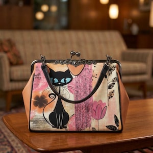 May include: A handbag featuring a black cat illustration with blue eyes, set against a background of pink and orange abstract shapes, floral motifs, and black leaves. The bag has a dark metal frame, a kiss-lock closure, and black handles.