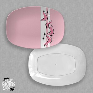 May include: Two oval serving platters. The top platter is pink with a white and gray stripe down the center, featuring pink boomerang shapes and black starbursts. The bottom platter is white.
