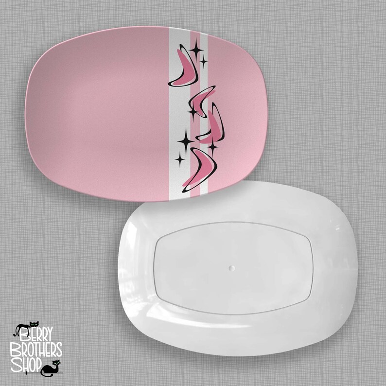 May include: Two oval serving platters. The top platter is pink with a white and gray stripe down the center, featuring pink boomerang shapes and black starbursts. The bottom platter is white.