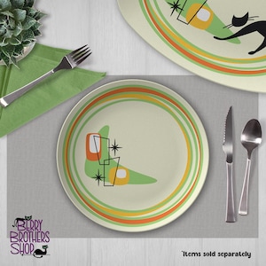 May include: A round ceramic plate featuring a cream-colored base with concentric orange and green circular rings. The design includes a stylized green boomerang shape, two orange rectangular accents, and black starburst icons. A fork, knife, and spoon are present.