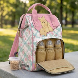 May include: A pink and light blue plaid backpack with a floral pattern and a Winnie the Pooh graphic on the front flap. The bag features a front compartment containing three baby bottles and a small clear container of cereal puffs beside it.