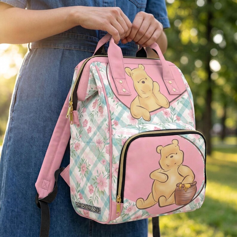 May include: A pink and light blue plaid backpack featuring floral patterns and illustrations of Winnie the Pooh. The bag has a top handle, adjustable shoulder straps, a front zippered pocket, and gold-toned hardware accents throughout the design.