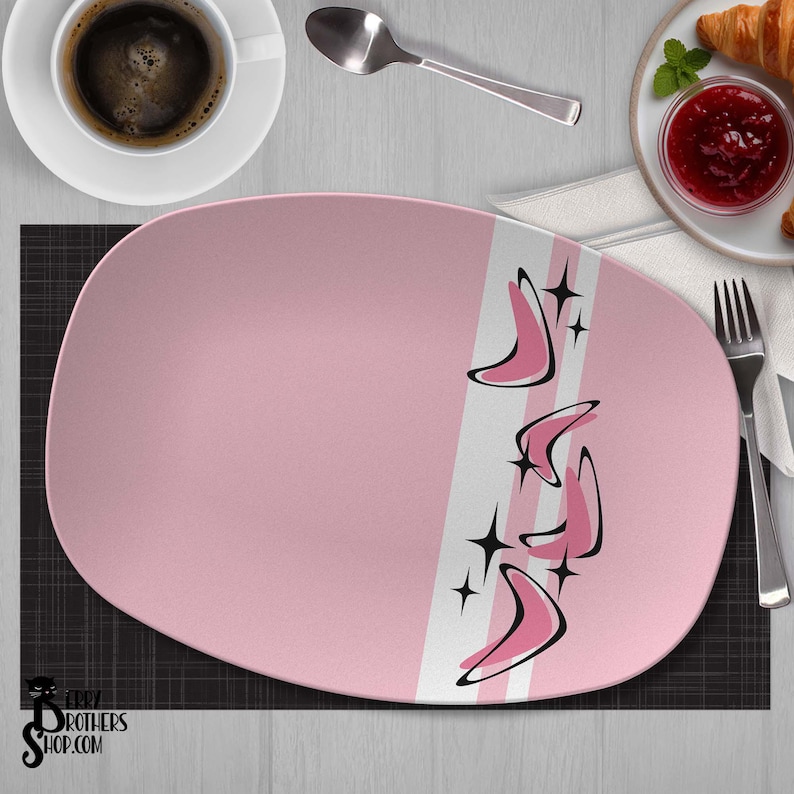 May include: An oval pink serving platter featuring a white vertical stripe and a pattern of black and pink boomerang shapes accented with four-pointed stars. The platter has a smooth finish and is centered on a dark gray textured surface.