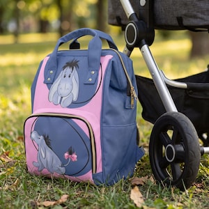 May include: A blue and pink backpack featuring Eeyore from Winnie the Pooh. The bag has a top handle, gold-tone zippers, and two circular graphic prints of the character. One print shows Eeyore's face, and the other shows the full body with a pink bow.