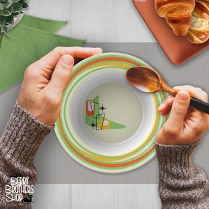May include: A round ceramic bowl featuring a light green center with a stylized boomerang graphic, orange and black geometric accents, and starburst details. The rim is decorated with concentric rings in shades of green, orange, and cream.