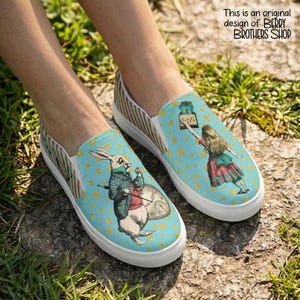 May include: A pair of blue slip-on canvas shoes with a whimsical Alice in Wonderland design. The shoes feature a blue background with gold polka dots and a print of Alice holding a bottle that says "Eat Me" on one and the white rabbit wearing a waistcoat and holding a pocket watch on the other shoe. The pattern also includes a crown and other details around the heel.