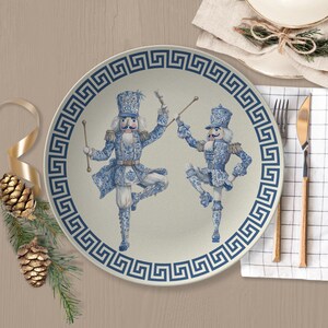 May include: A round ceramic dinner plate featuring a blue and white chinoiserie-style pattern of two nutcracker figures in dancing poses. The plate is bordered by a dark blue Greek key motif on a light cream background, creating a festive design.