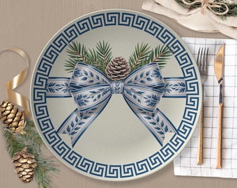 May include: A round ceramic dinner plate featuring a central illustration of a blue and white patterned ribbon tied in a bow, accented with green pine needles and a brown pinecone. The plate has a dark blue Greek key border design.