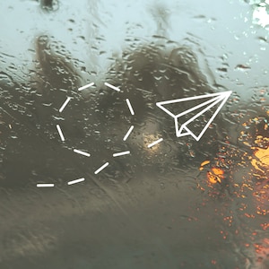 May include: A white paper airplane flying through a blurry background of raindrops on a window. A trail of dashes follows the airplane.