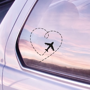 May include: A black airplane silhouette flying in a dashed line heart shape on a car window. The background is a blurry pink and purple sunset.